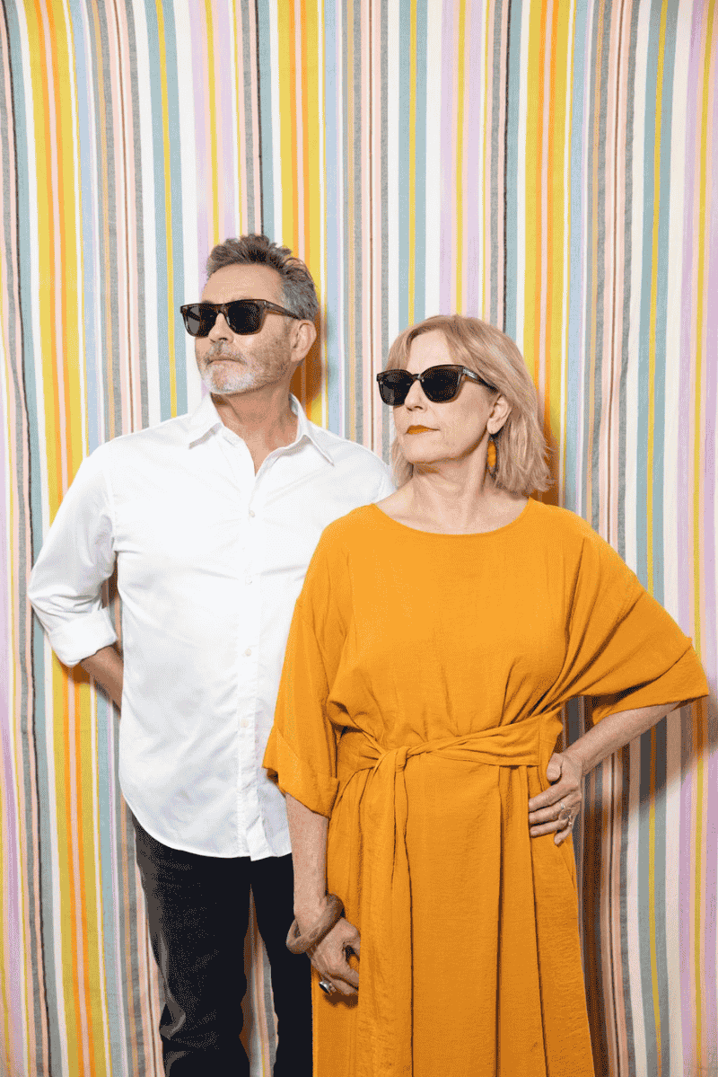 Two people, a man and woman, standing against a colorful striped background wearing vital sunglasses